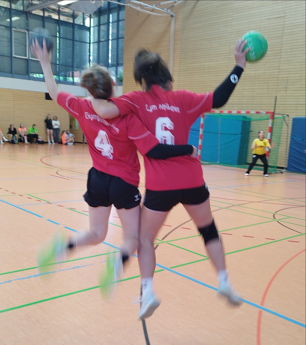 Auf dem Bild sind zwei Handballerinnen im Sprung. Im Hintergrund ist ein Tor und die Torwärtin zu sehen.