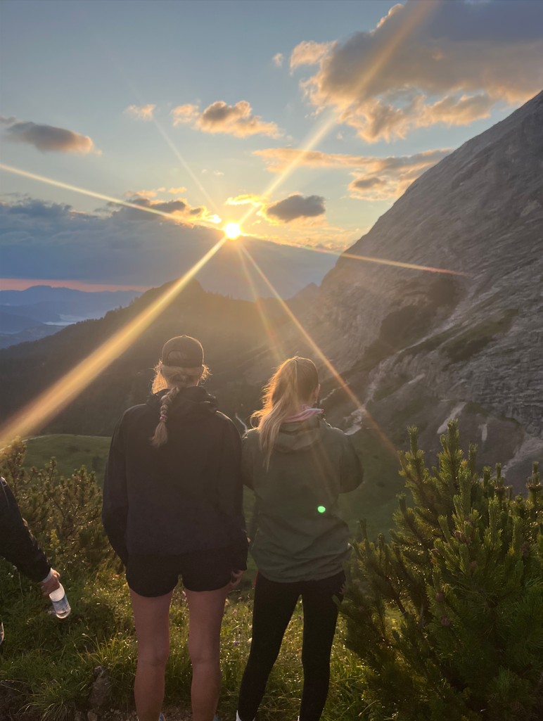 Zu sehen sind zwei Schülerinnen beim Sonnenaufgang in den Alpen.