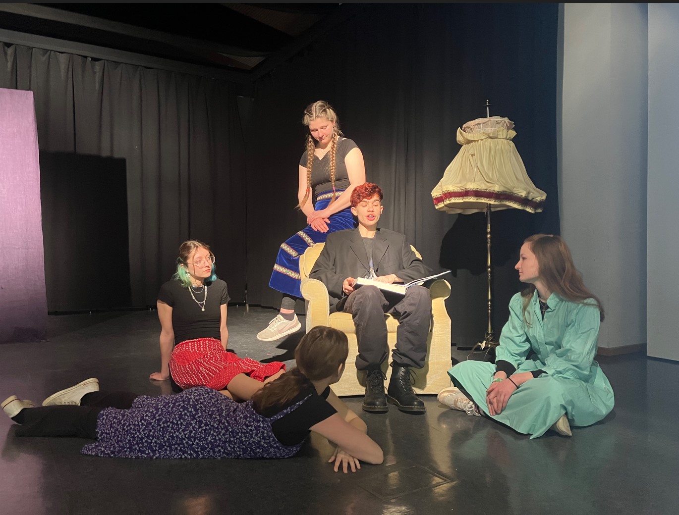 Fünf Schülerinnen und Schüler spielen auf der Bühne Theater. Im Zentrum des Bildes sitzt ein Erzähler in einem gelben Sessel und ließt aus einem Märchenbuch vor.