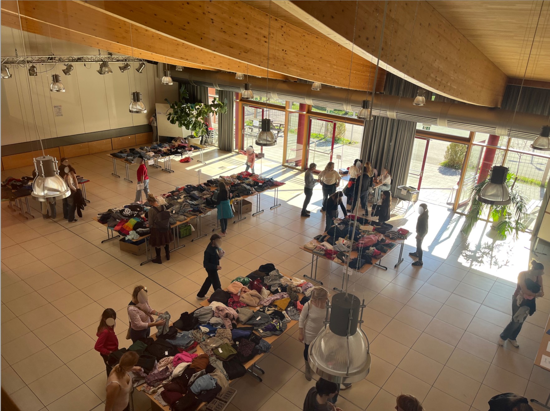 Zu sehen ist die Schulaula. Auf Schultischen, die zu Marktständen umgewandelt wurden, ist Kleidung drapiert, durch welche die Besucherinnen und Besucher stöbern.